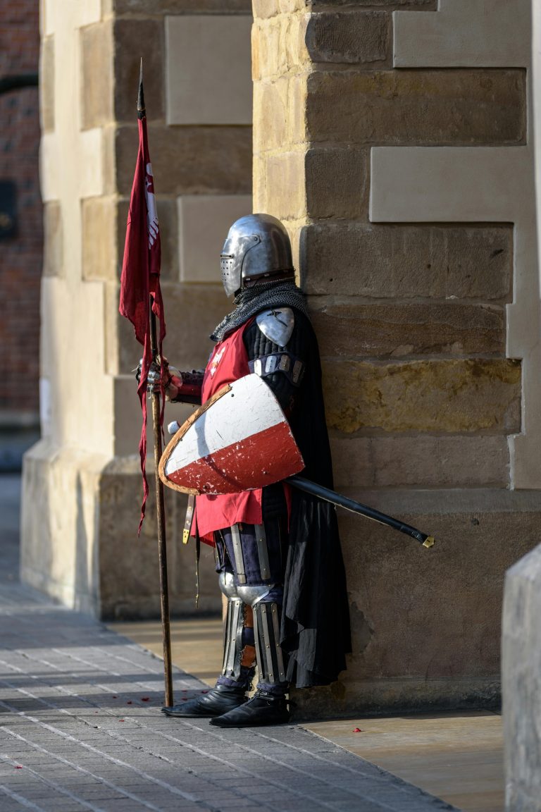 A medieval knight in armor stands guard with a shield and flag outside a stone building.