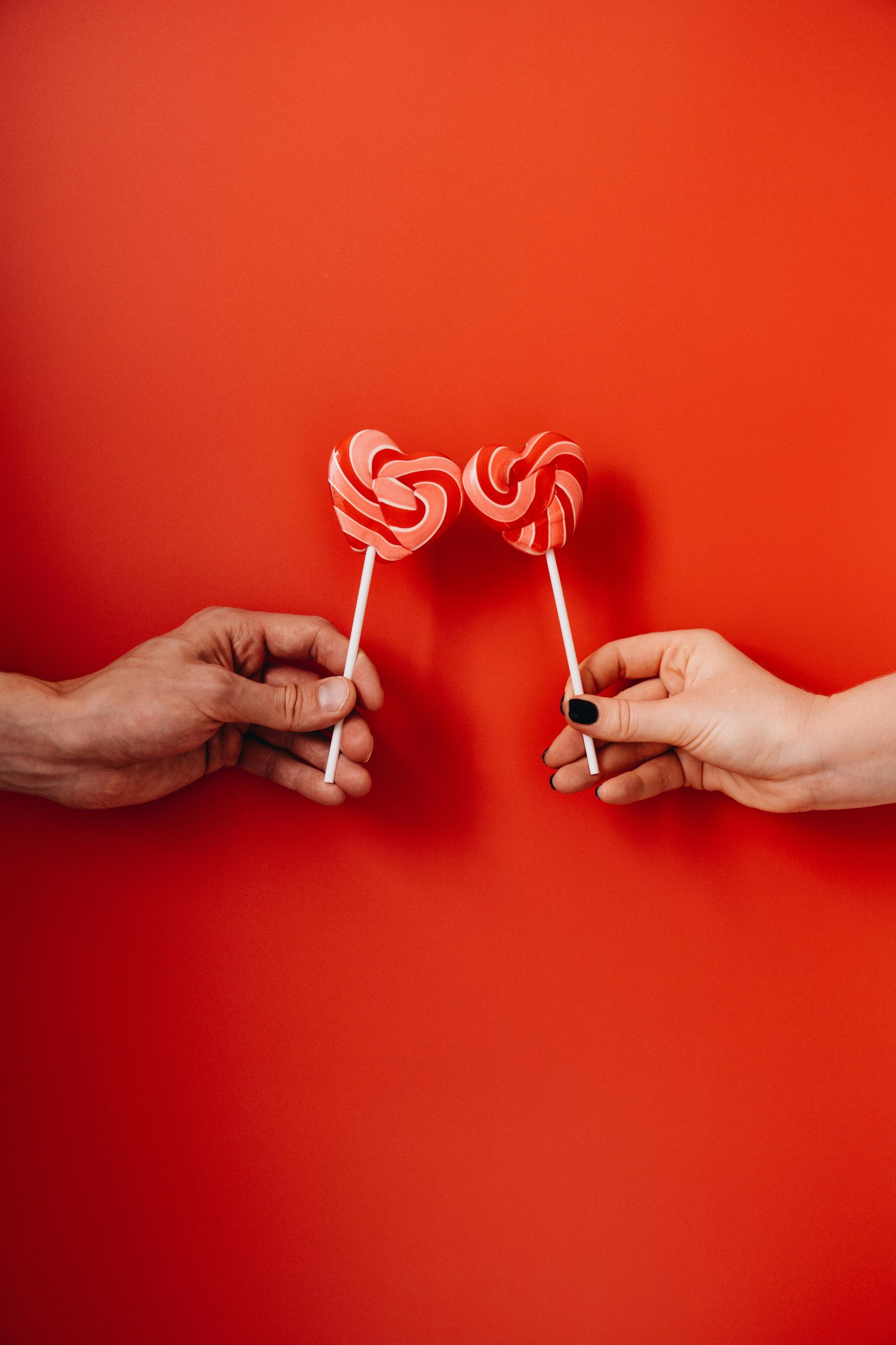 Two hands holding heart-shaped lollipops against a vibrant red background, symbolizing love.